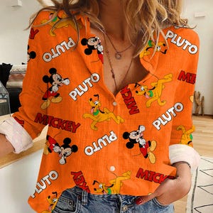 May include: Orange button-down shirt featuring Mickey Mouse and Pluto cartoon characters. The shirt has long sleeves with white cuffs and is patterned with the words "Mickey" and "Pluto". The shirt is made of a lightweight fabric.
