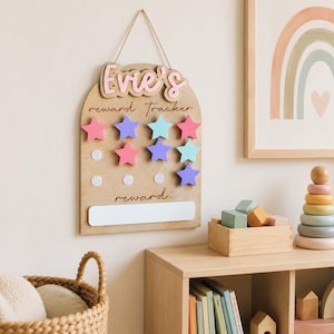 Może przedstawiać: Drewniana tablica nagr&oacute;d z imieniem "Evie's" w r&oacute;żowych literach. Tablica ma szereg kolorowych magnes&oacute;w w kształcie gwiazd w kolorze r&oacute;żowym, niebieskim i turkusowym. Na tablicy wydrukowano słowa "reward tracker" i "reward:"