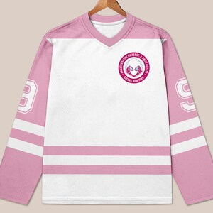 May include: A white and pink hockey jersey with long sleeves. The jersey features a V-neck collar and the number 99 on the sleeves. A circular emblem with text and a spider-themed graphic is on the chest.