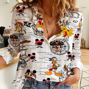 May include: A white button-down shirt with a long sleeve featuring a cartoon print of Mickey Mouse, Donald Duck, and other Disney characters. The shirt has a relaxed fit and a classic collar.