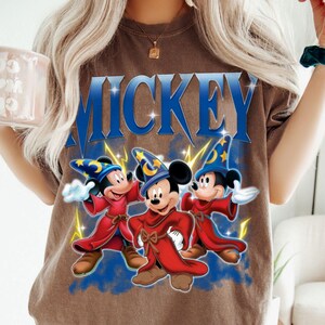 Puede incluir: Camiseta marr&oacute;n con la palabra "MICKEY" en letras azules. La imagen muestra tres personajes de Mickey Mouse vestidos con t&uacute;nicas rojas y sombreros de mago azules con estrellas amarillas, sobre un fondo azul.