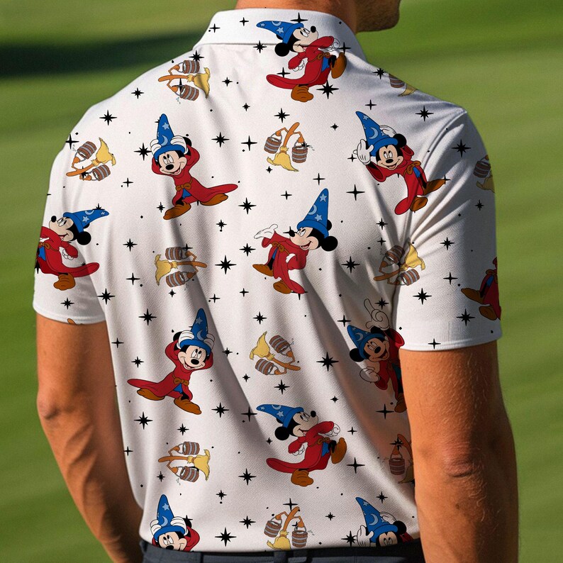 Puede incluir: Polo blanco con un estampado repetido de Mickey Mouse con un disfraz de mago, escobas y estrellas. El personaje de dibujos animados viste un sombrero de mago azul y una t&uacute;nica roja. La camisa tiene mangas cortas y cuello.