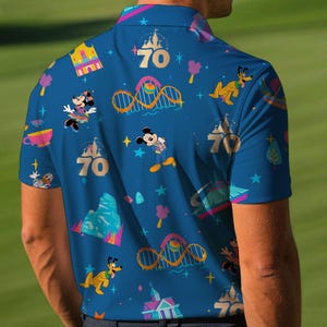 May include: Blue polo shirt featuring a colorful pattern with cartoon characters, roller coasters, and the number 70. The shirt has a button-down collar and short sleeves. The design includes Mickey Mouse, Minnie Mouse, and Pluto.