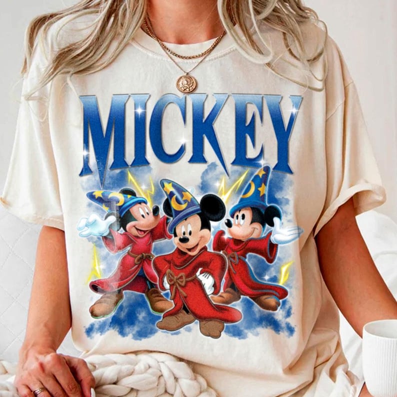 Puede incluir: Camiseta color crema con la palabra "MICKEY" en azul, sobre un gr&aacute;fico de tres personajes de Mickey Mouse vestidos como hechiceros con t&uacute;nicas rojas y sombreros de mago azules. El dise&ntilde;o incluye rayos y acentos de nubes azules.