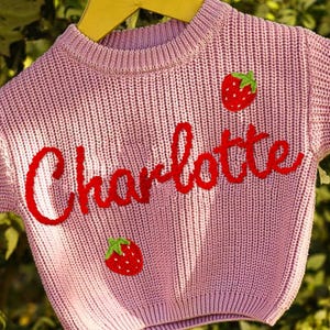 May include: A light pink knit sweater with the name "Charlotte" in red script across the front. Two red strawberry patches with green leaves are also on the sweater. The sweater is hanging on a yellow hanger.