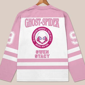 May include: Pink and white Ghost-Spider hockey jersey. The jersey features the text "GHOST-SPIDER" above a circular emblem with a spider-themed mask and the text "AN ALTERNATIVE UNIVERSE, A TWIST OF FATE, QUEENS, NEW YORK". The name "GWEN STACY" is below the emblem.
