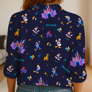 May include: Navy blue button-up shirt featuring a Disneyland theme. The shirt is adorned with colorful illustrations of Mickey Mouse, Donald Duck, and other Disney characters, along with the text "Disneyland" and the number "70".