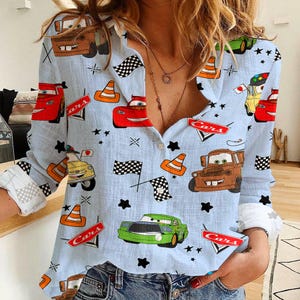 May include: Light blue button-down shirt featuring a repeating pattern of cartoon cars, traffic cones, checkered flags, and stars. The long-sleeved shirt has white cuffs and a classic collar. The word "Cars" is printed on red signs throughout the design.
