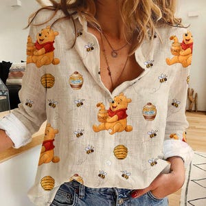 May include: Beige button-down shirt featuring a repeating pattern of Winnie the Pooh, honey pots, bees, and beehives. The long-sleeved shirt has white cuffs and a relaxed fit. The shirt is made of a lightweight fabric.