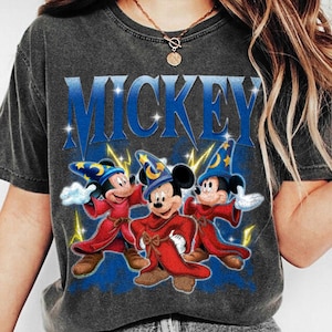 Puede incluir: Camiseta gris oscuro con la palabra "MICKEY" en azul, con tres personajes de dibujos animados de Mickey Mouse vestidos de magos. Los personajes visten t&uacute;nicas rojas y sombreros de mago azules con estrellas doradas. Rayos amarillos y estrellas rodean a los personajes.