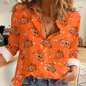May include: Orange button-down shirt with a repeating pattern of cartoon clownfish. The long-sleeved shirt has white cuffs and a relaxed fit. The clownfish are orange, white, and black, with smiling faces. The background has white dots and star shapes.