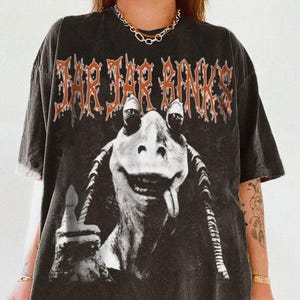 May include: Black t-shirt with a graphic print of Jar Jar Binks from Star Wars. The text "Jar Jar Binks" is printed in orange above the image.