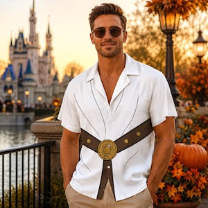 May include: A white short-sleeved button-down shirt featuring a printed brown belt design across the midsection with a central gold sun emblem. The shirt has a notched collar and a lightweight fabric construction with thin black line details.