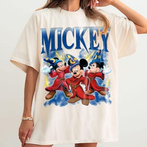 Puede incluir: Camiseta color crema con la palabra "MICKEY" en letras azules en bloque sobre un gr&aacute;fico de tres personajes de Mickey Mouse vestidos como hechiceros con t&uacute;nicas rojas y sombreros de mago. El dise&ntilde;o incluye rayos amarillos y un fondo azul.
