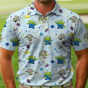 May include: A light blue short-sleeve polo shirt featuring an all-over repeating pattern of Buzz Lightyear and green three-eyed aliens from Toy Story, interspersed with yellow planets, blue Saturn-like rings, and small blue and green starbursts.