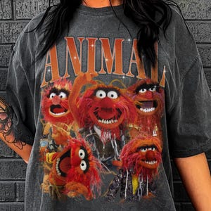 May include: Dark gray t-shirt featuring the word "ANIMAL" in orange letters above a graphic of the Muppets' Animal and his bandmates. The image shows the characters with red fur and expressive faces.