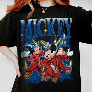 Puede incluir: Camiseta negra con la palabra "MICKEY" en azul y tres personajes de Mickey Mouse vestidos de magos. Llevan sombreros de mago azules y t&uacute;nicas rojas, con rayos amarillos. Estilo de dibujos animados.