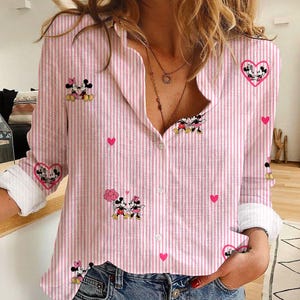 May include: Pink and white striped button-down shirt featuring Mickey and Minnie Mouse graphics and pink hearts. The long-sleeved shirt has white cuffs and a classic collar. The shirt is made of a lightweight fabric.