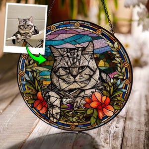 May include: Round stained glass panel featuring a gray tabby cat with a stern expression, surrounded by colorful floral and foliage elements. The panel has a decorative border and is suspended by a chain. The image also includes a smaller photo of the cat.