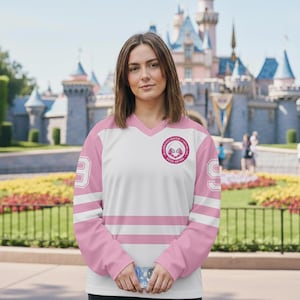 May include: White and pink hockey-style jersey with a V-neck and long sleeves. The jersey features pink stripes across the chest and sleeves, with the number 9 on the sleeves. A circular emblem is on the chest.
