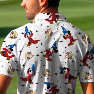 Puede incluir: Polo blanco con un estampado repetido de Mickey Mouse con un disfraz de mago, estrellas y otros elementos de dibujos animados. La camisa tiene mangas cortas y un cuello cl&aacute;sico. El dise&ntilde;o incluye colores rojo, azul y amarillo.