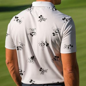 Puede incluir: Un polo de color claro con un patr&oacute;n repetido de ilustraciones de Mickey Mouse en blanco y negro. La camisa de manga corta tiene un cuello cl&aacute;sico. La persona lleva la camisa sobre un fondo verde.