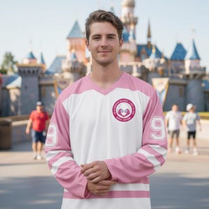 May include: A white and pink hockey-style jersey with the number 9 on the sleeves. The jersey has a V-neck and a circular emblem with text and a graphic. The sleeves have two white stripes. The jersey is worn by a person.