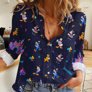 May include: Navy blue button-down shirt featuring Disney characters like Mickey Mouse, Donald Duck, and Minnie Mouse. The shirt has a colorful castle graphic and the word "Disneyland". The long sleeves have white cuffs.