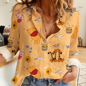 May include: A button-down, long-sleeve shirt in a light yellow color, featuring cartoon illustrations of Winnie the Pooh, Piglet, and Tigger. The shirt has a V-neck and white cuffs. Honey pots, butterflies, and paper airplanes are also printed on the shirt.