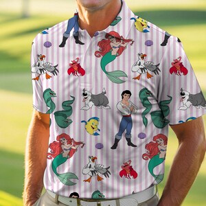 May include: A short-sleeved polo shirt featuring a repeating pattern of characters from The Little Mermaid, including Ariel, Prince Eric, Flounder, Sebastian, Scuttle, and Max, set against a background of vertical white and light pink stripes.