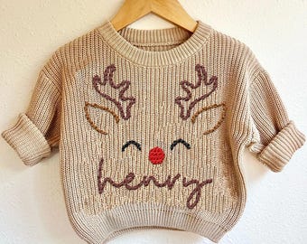 May include: Tan knit sweater with a reindeer face design. The sweater features embroidered antlers, eyes, and a red nose. The name "Henry" is embroidered below the face. The sweater is hanging on a wooden hanger.