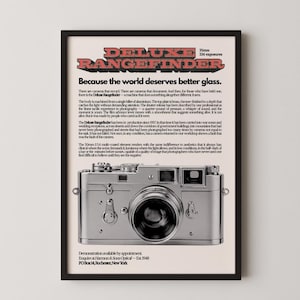 Op de afbeelding: Een ingelijste advertentie voor een Deluxe Rangefinder camera. De poster toont de camera in het midden, met tekst erboven en eronder. De tekst luidt: "Omdat de wereld beter glas verdient."