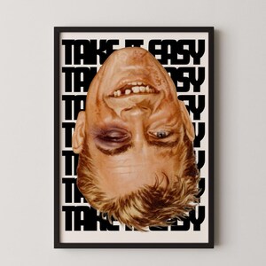 May include: A framed art print featuring a portrait of a man with a black eye and a smiling expression. The words "TAKE IT EASY" are printed in a repeating pattern in black and white.