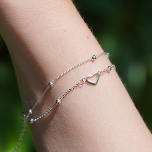 Pode incluir: Duas pulseiras de corrente prateadas com pequenas contas e um pingente em forma de coração. As pulseiras estão sobrepostas no pulso de uma pessoa.