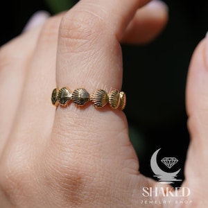 May include: Gold ring featuring a design of multiple seashells. The ring is worn on a person's finger.