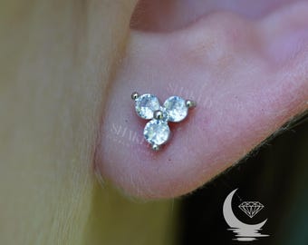 Puede incluir: Primer plano de un pendiente de plata con tres gemas redondas y transparentes dispuestas en un racimo triangular. El pendiente está colocado sobre la piel de una oreja. El logotipo de la tienda "SHAKED JEWELRY SHOP" es visible.