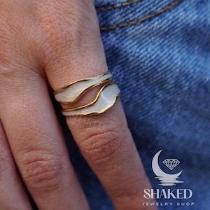 May include: A white and gold ring with a wavy design. The ring is on a finger wearing a blue denim shirt.