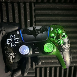 May include: Custom black and green video game controller with Batman and Joker graphics. The controller has illuminated buttons and a Batman logo on the touchpad. The design includes a green and purple colour scheme.