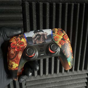 Könnte beinhalten: Angepasster schwarzer Videospiel-Controller mit orangefarbenen und roten Akzenten. Der Controller hat ein zentrales Bild und flammenartige Muster an den Seiten. Die Tasten sind ebenfalls orange und rot.