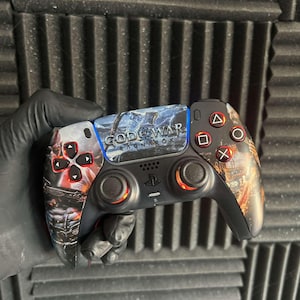 May include: A custom PlayStation 5 controller featuring artwork from the God of War Ragnarok video game. The controller has a black base with red, blue, and orange accents. The face buttons are red, and the touchpad displays the game's title.