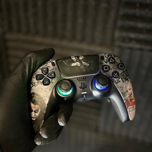 Könnte beinhalten: Angepasster schwarz-grauer Videospiel-Controller mit Joker-Bildern. Der Controller hat grün und blau beleuchtete Joysticks und verschiedene Tastenmuster. Der Controller wird von einer behandschuhten Hand gehalten.