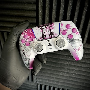 Könnte beinhalten: Ein weiß-rosa Custom-Videospiel-Controller mit japanisch inspiriertem Design. Der Controller zeigt eine Blumen- und Berglandschaft mit dem Text "ゆきな". Die Knöpfe sind rosa, und der Controller wird von einer behandschuhten Hand gehalten.