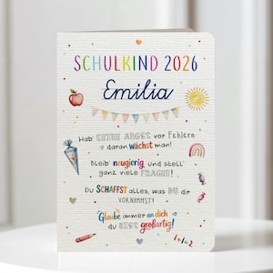 Könnte beinhalten: Eine weiße Grußkarte mit bunten Illustrationen und Text. Die Karte trägt die Aufschrift "SCHULKIND 2026 Emilia" und enthält eine Zeichnung eines Apfels, einer Sonne, eines Regenbogens und andere kleine Illustrationen. Die Karte hat deutschen Text.