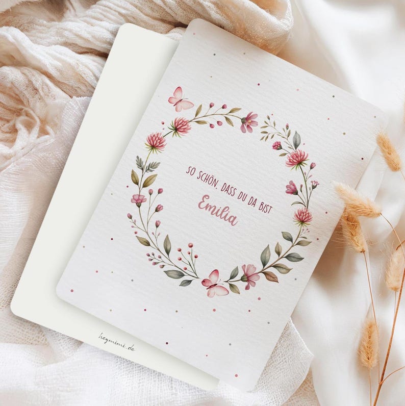 May include: A greeting card with a floral wreath design in pink and green, featuring the name "Emilia" in pink script. The card includes the text "SO SCH&Ouml;N, DASS DU DA BIST" and a small butterfly illustration. The card is on a white background.