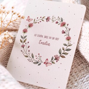 May include: A greeting card with a floral wreath design. The wreath features pink flowers, green leaves, and butterflies. The card has the text "SO SCH&Ouml;N, DASS DU DA BIST Emilia" in a script font. The card is on a soft, textured background.