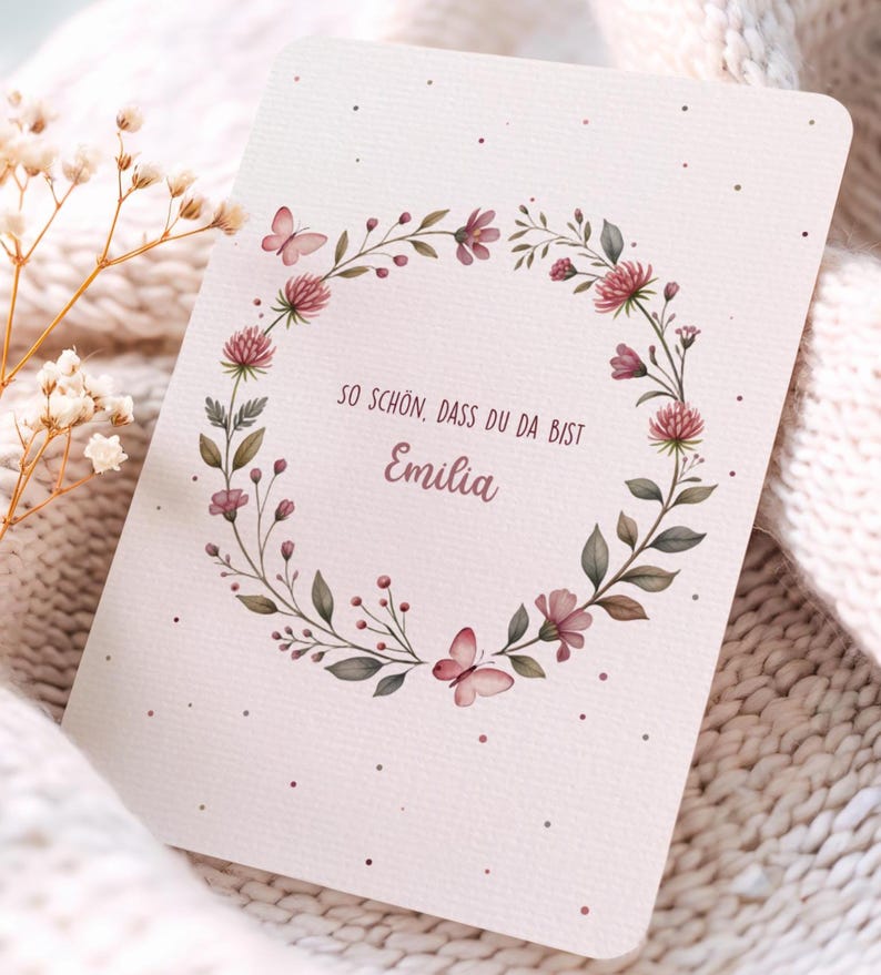 May include: A greeting card with a floral wreath design. The wreath features pink flowers, green leaves, and butterflies. The card has the text "SO SCH&Ouml;N, DASS DU DA BIST Emilia" in a script font. The card is on a soft, textured background.