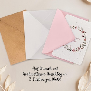 May include: A greeting card with a floral wreath design, nestled in a pink envelope, with a white and brown envelope. The card has text in a script font. The text on the image says "Auf Wunsch mit hochwertigem Umschlag in 3 Farben zur Wahl."