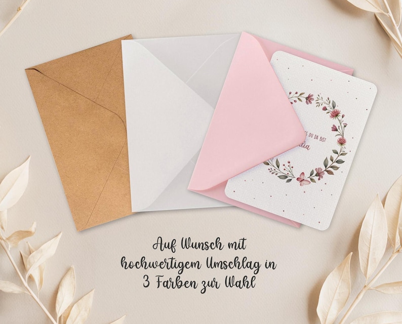 May include: A greeting card with a floral wreath design, nestled in a pink envelope, with a white and brown envelope. The card has text in a script font. The text on the image says "Auf Wunsch mit hochwertigem Umschlag in 3 Farben zur Wahl."