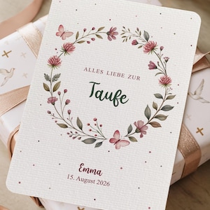 Puede incluir: Una tarjeta crema con un dise&ntilde;o de corona floral, con flores rosas y mariposas. El texto "ALLES LIEBE ZUR Taufe" est&aacute; centrado, con el nombre "Emma" y la fecha "15. August 2026" debajo. La tarjeta est&aacute; sobre un regalo envuelto.