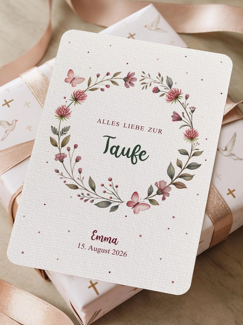 Puede incluir: Una tarjeta crema con un dise&ntilde;o de corona floral, con flores rosas y mariposas. El texto "ALLES LIEBE ZUR Taufe" est&aacute; centrado, con el nombre "Emma" y la fecha "15. August 2026" debajo. La tarjeta est&aacute; sobre un regalo envuelto.
